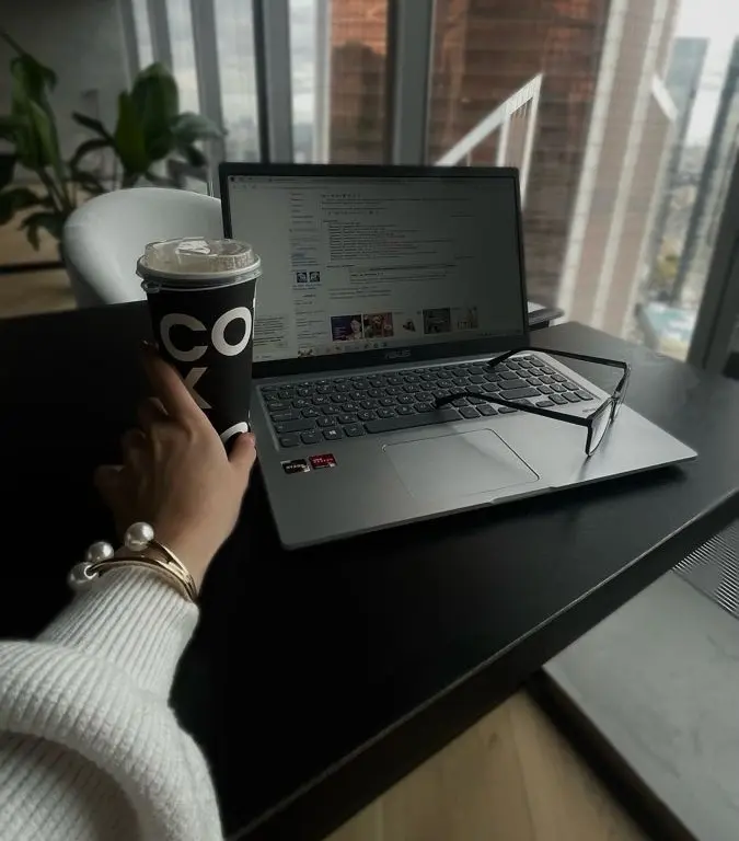 Hand holding a coffee cup beside an open laptop with glasses on the keyboard in a high-rise workspace.