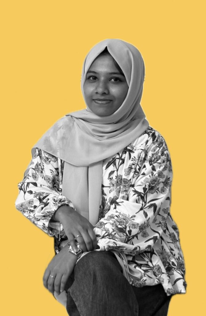 Person wearing a floral-patterned top and a light-colored hijab, seated against a solid yellow background.